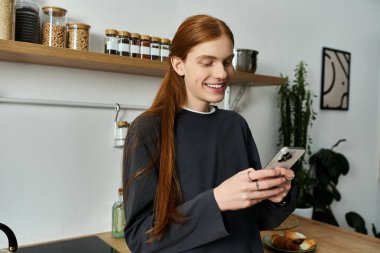 A good looking redhead in a sweatshirt smiles while using his phone in a cozy apartment. clipart