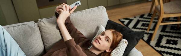 Charming redhead young man lies comfortably on a sofa, capturing a moment on his phone, banner