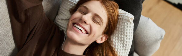 A cheerful young man with long red hair lies comfortably on a couch, embracing a relaxed moment, banner