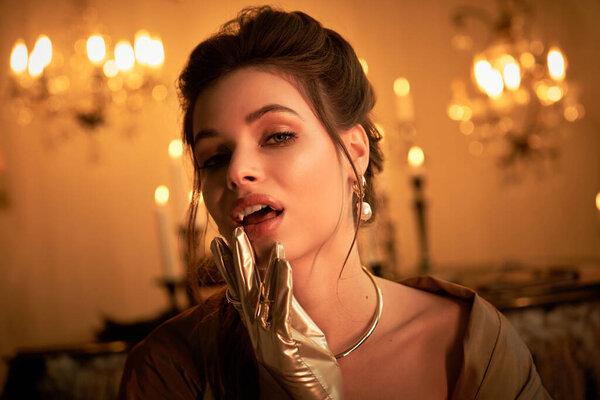 A young vampire touches her lips, cloaked in candlelight, exuding mystery.