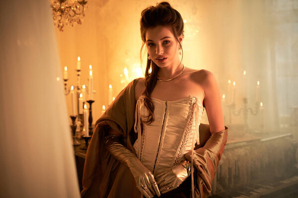 Уверенная в себе молодая женщина в элегантной одежде стоит посреди мерцающих свечей, вызывая мистическую атмосферу.