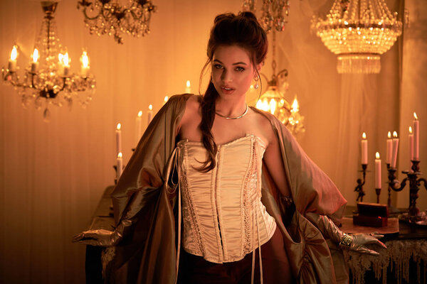 A young woman in elegant attire stands in a candle lit mystical room, radiating enchantment.
