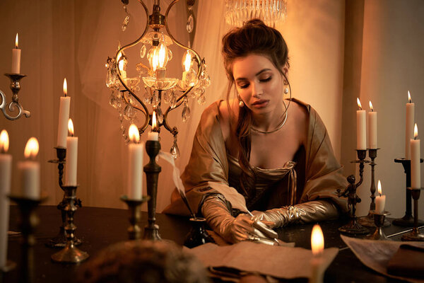 Красивая молодая женщина, одетая в фантастический наряд, пишет в окружении свечей и теней..