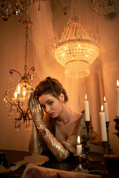 Молодая женщина в золотых перчатках сидит в тускло освещенной комнате с мерцающими свечами.