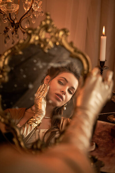 A young woman in fantasy attire admires her reflection in a candlelit, ornate mirror.