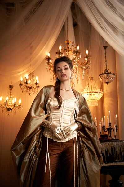 A stunning young woman in a vampire outfit poses elegantly in a candlelit room.
