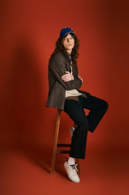 Şık genç bir adam, cesur kırmızı bir arka planda, bir taburede rahatça oturuyor, kendine güveni ve tarzı var..