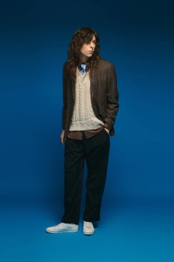 Şık bir genç adam, parlak mavi bir arka plana karşı katmanlı giysiler içinde güvenle duruyor..