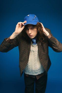 Young man showcases a brown blazer and blue cap against a vibrant blue background. clipart