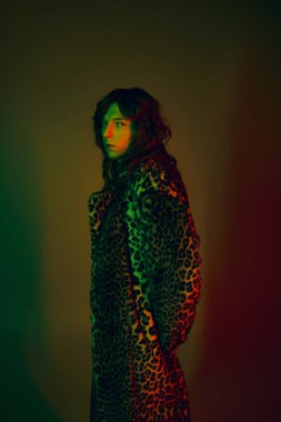 Şık bir genç adam kendine güvenerek duruyor, renkli ışıkların altında şık bir leopar desenli ceket giyiyor..