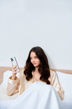 A beautiful young woman sits comfortably in bed, absorbed in her phone, surrounded by cozy linens. clipart