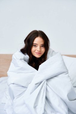 A young woman enjoys a serene moment, wrapped snugly in a blanket while sitting on a bed. clipart