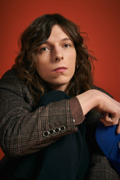 Опрятный молодой человек с волнистыми волосами уверенно сидит, демонстрируя свой шикарный наряд и модный стиль.