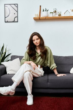 A lovely young woman poses elegantly on a dark sofa surrounded by modern decor and plants. clipart