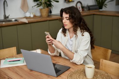 Güzel genç bir kadın mutfak masasında otururken akıllı telefonuyla etkileşime giriyor..