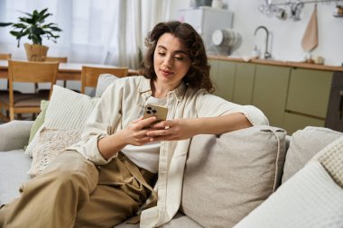 Rahat genç bir kadın rahat bir şekilde bir koltukta oturur ve sıcak bir evde akıllı telefonuyla meşgul olur..