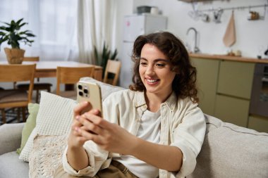 Güzel genç bir kadın, rahat bir evde akıllı telefonuyla meşgul olurken gülümsüyor..