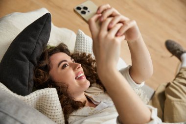 Güzel genç bir kadın rahat bir kanepede uzanıyor, mutlu bir şekilde akıllı telefonuna bakıyor..