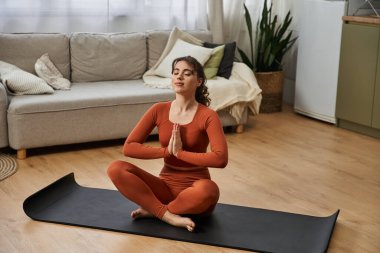 A beautiful young woman enjoys a moment of mindfulness while sitting peacefully on a mat at home. clipart