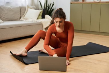 Pratik yapmaya odaklanmış, güzel bir genç kadın laptopunu evde yoga için kullanıyor..