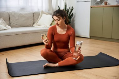 Güzel genç bir kadın evde yoga minderinde oturup suyunu yudumluyor ve telefonundan kayıyor..