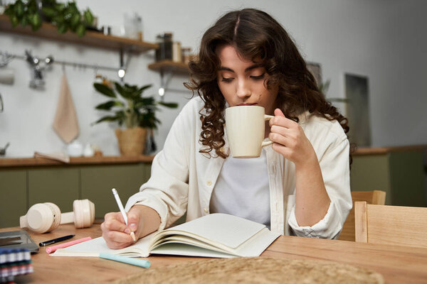 Beautiful young woman relaxes at home, sipping a warm drink while writing in her notebook.