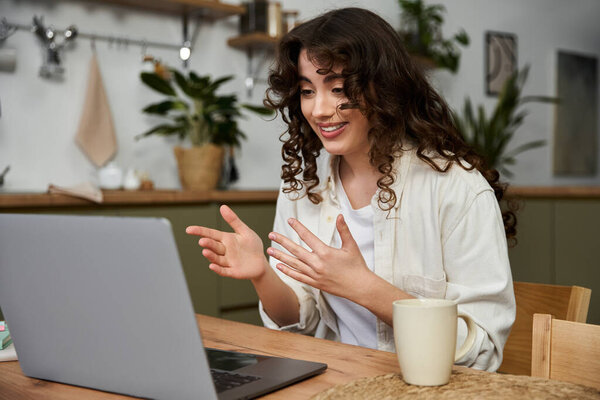 Улыбающаяся молодая женщина волнующе взаимодействует во время видеозвонка дома с горячим напитком поблизости.