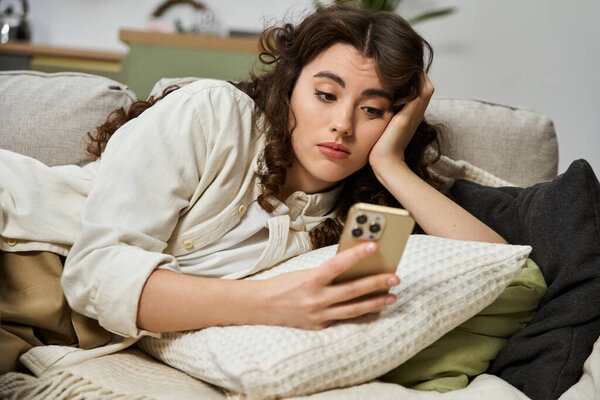 A beautiful young woman relaxes at home, engrossed in her phone while resting on a cushy sofa.