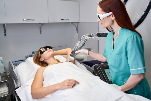 A technician conducts laser hair removal on a young woman in a clean clinic.