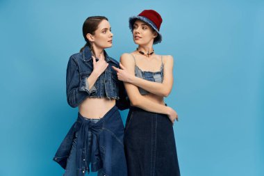 İki şık kotlu kız kardeş neşeli bir şekilde parlak mavi bir arkaplanda poz veriyorlar..