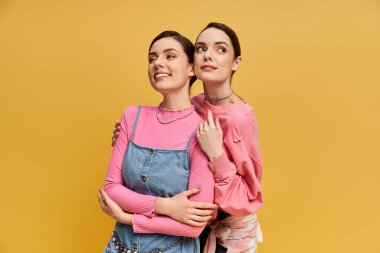 İkiz kardeşler neşeli bir anı paylaşıyorlar, parlak sarı bir arkaplanın önünde ışıldıyorlar..