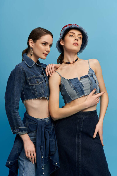 Two beautiful sisters in trendy denim outfits pose playfully against a blue background.
