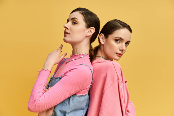 Две прекрасные сестры-близнецы стоят спиной к спине, излучая уверенность и очарование в яркой обстановке.