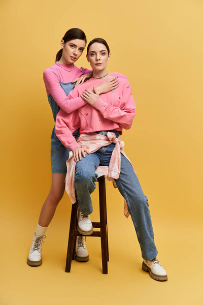 Two beautiful twin sisters sit together, sharing a warm moment in stylish outfits and playful poses.