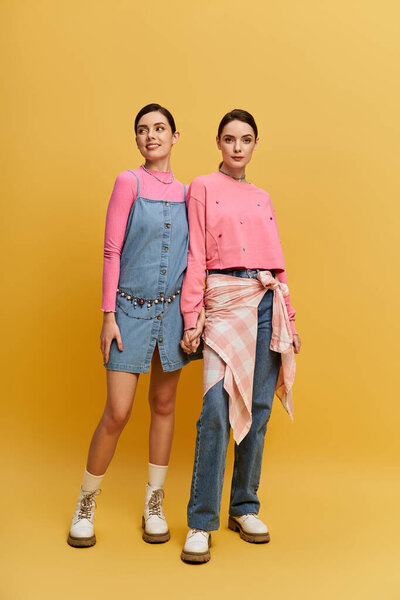 Two stylish sisters hold hands against a bright yellow backdrop.