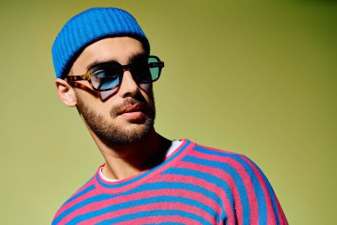 Tarz sahibi genç bir adam yeşil bir stüdyoda kendine güvenerek poz veriyor. Moda kıyafetlerini ve gözlüklerini sergiliyor..