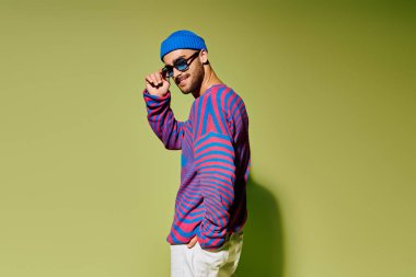 Genç bir adam şık kıyafetini ve parlak güneş gözlüklerini yeşil bir zemine karşı sergiliyor..