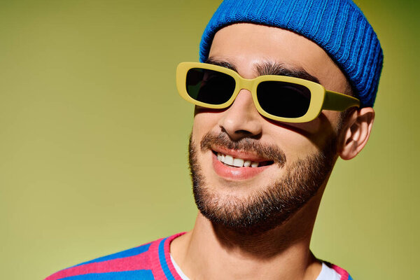 A young man in stylish attire and vivid sunglasses flaunts his bold fashion sense in a studio.