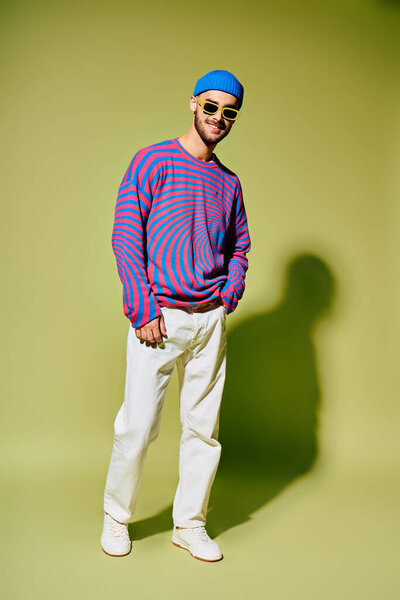 A young man confidently displays his trendy outfit and vibrant sunglasses in a studio setting.