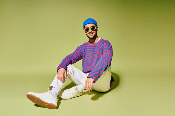 A confident young man lounges in trendy attire and sunglasses against a bright green backdrop.