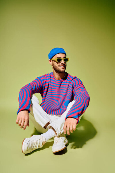 Handsome young man sits casually, modeling a vibrant outfit against a green backdrop.