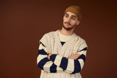 Yakışıklı, şık giyinmiş ve bereli bir genç, kahverengi bir arka plana karşı güvenle poz veriyor..