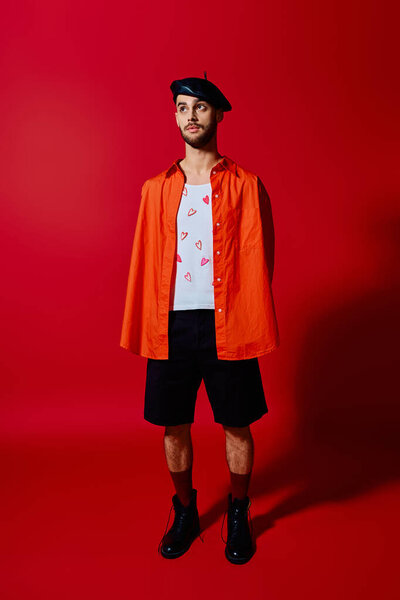 Молодой человек в узорчатой футболке и берете позирует на ярком красном фоне.