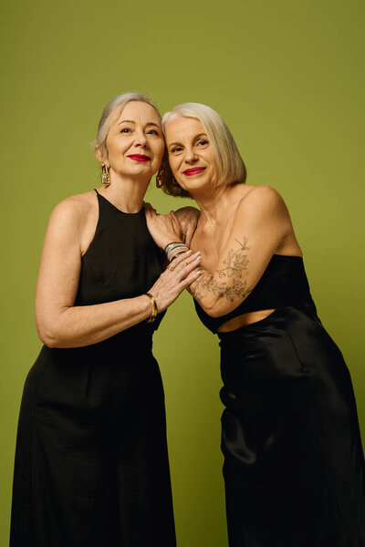 Две зрелые женщины разделяют стильные объятия, излучающие любовь и уверенность на ярком фоне.