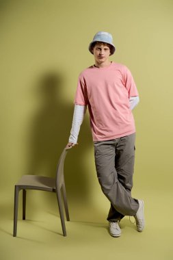 Pembe tişört ve günlük pantolon giyen genç bir adam, şık bir sandalyeye yaslanıyor..