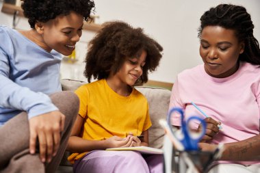 Three family members share a joyful moment as they create art together on a cozy couch. clipart