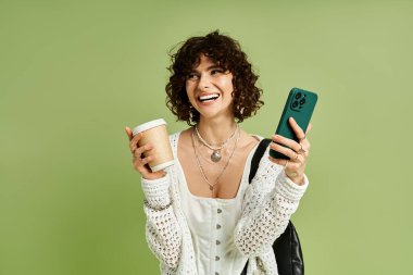 Neşeli bir kadın elinde kahve fincanı ve akıllı telefonuyla gülüyor. Parlak yeşil bir alana kurulmuş..