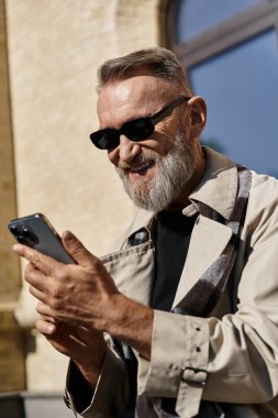 Ceket ve güneş gözlüğü takmış kıdemli bir adam şehir manzarasında yürür ve telefona bakar..