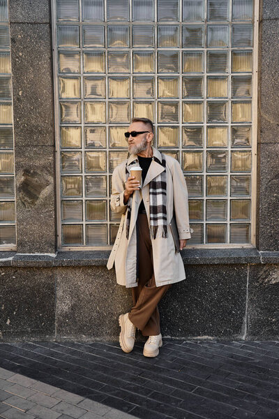 A dapper senior man in trendy attire stands against a chic glass wall, holding a coffee cup.