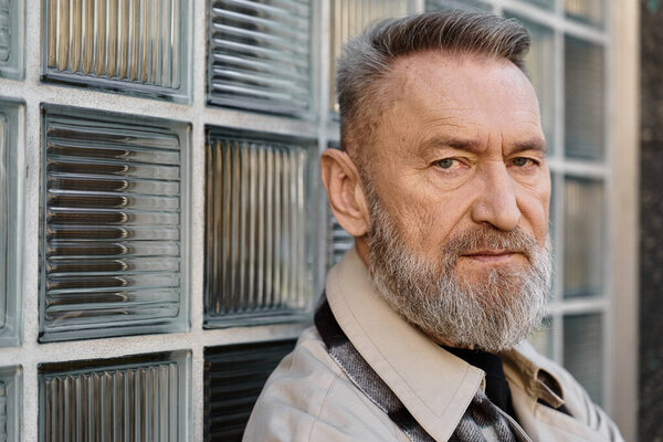 A senior man stands against a glass wall, exuding confidence and charm while in trendy clothing.